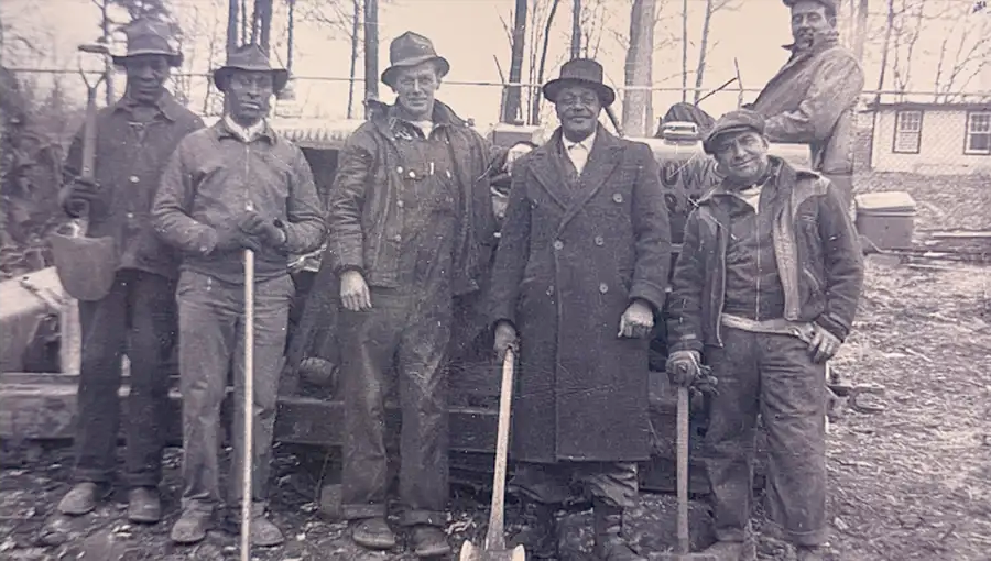 Founding-era Grabowski crew portrait — five men in long coats with shovels, in front of a small structure.