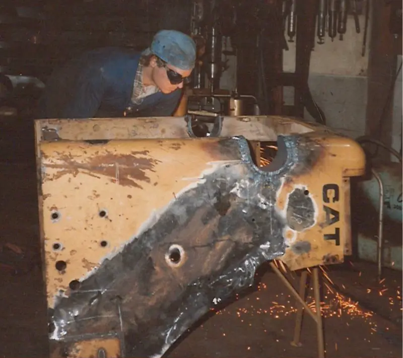 Worker welding the underside of a CAT excavator boom, sparks falling.